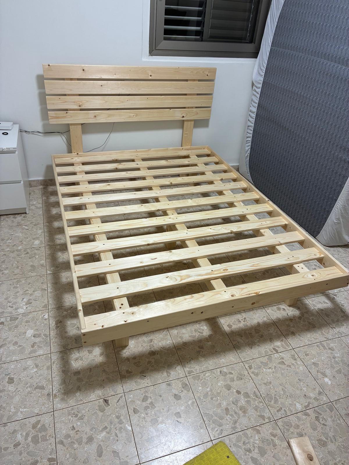 בסיס מיטה מוגבה מעץ אורן עם משענת ראש, לצד מזרן הנשען על הקיר